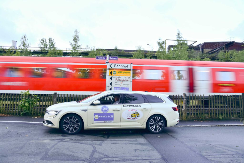City-Taxi Mietwagen fährt an einer S-Bahn am Bahnhof vorbei, im Hintergrund ein Wegweiser